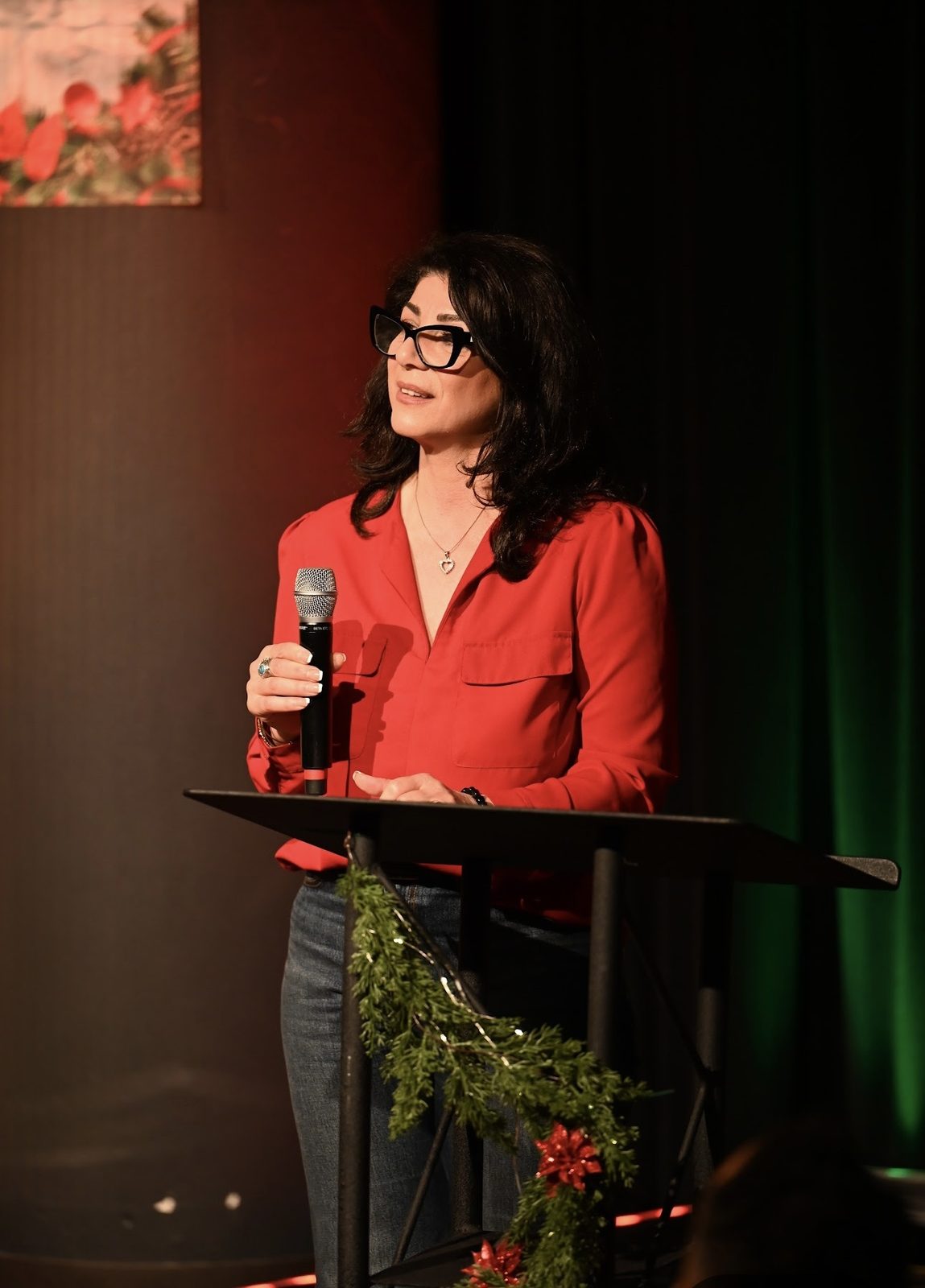 Kobi Julian speaking at a lectern with a microphone in a red blouse