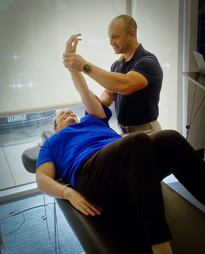 A man and woman appear to be playing or laughing together, with the woman lying on a treatment table and the man standing beside her, gesturing with their hands in a friendly manner.