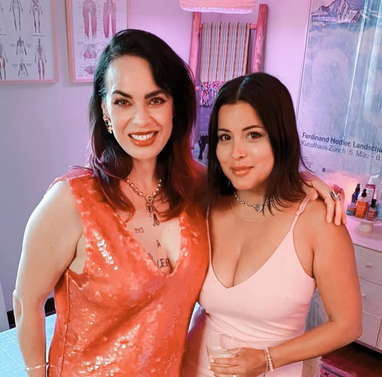 Two women smiling and posing together at a social event or party in an indoor setting, with a pink-toned background and posters on the wall.