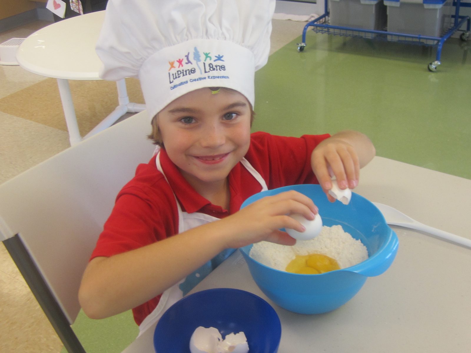 A Lupine Lane student in a chef's hat and apron cracking an egg into a bowl of flour