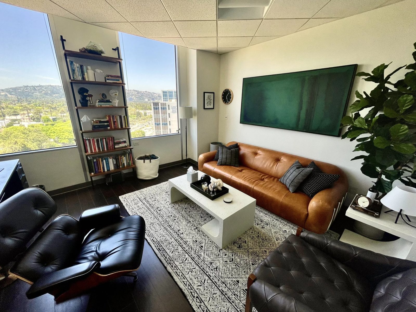 Modern living room with large windows, leather sofa, area rug, white coffee table, bookshelf, and green wall art.