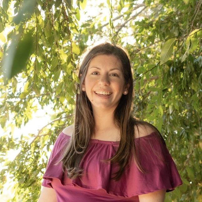Maggie Ochoa, founder of Coastal Kids Playspace, smiling in an off-the-shoulder magenta top beneath green leaves