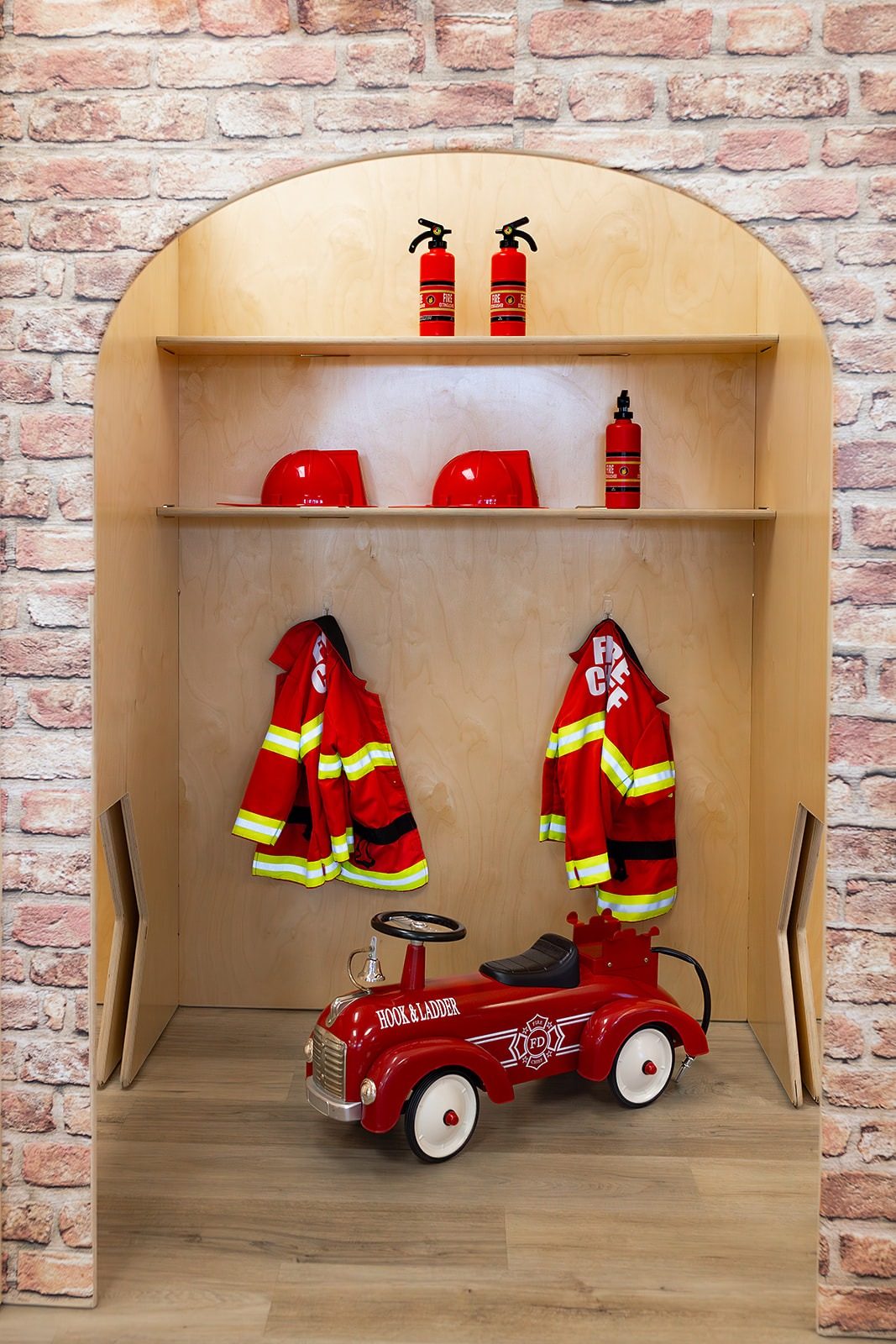 A firefighter dress-up nook at Coastal Kids Playspace with child-sized red jackets, helmets and a red ride-on fire truck under a wooden arch