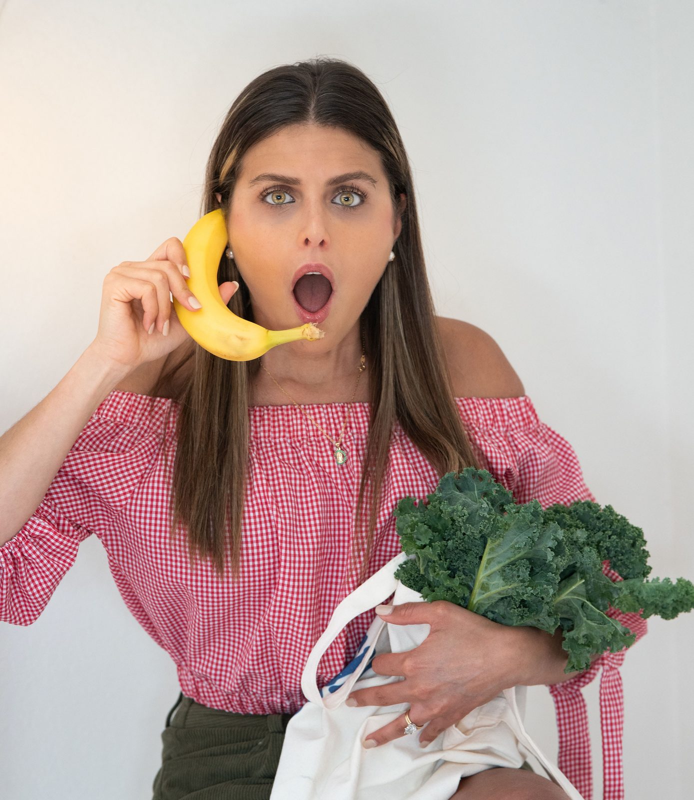 Maria Harrison in a red gingham top holding a banana to her ear like a phone and a tote bag of kale