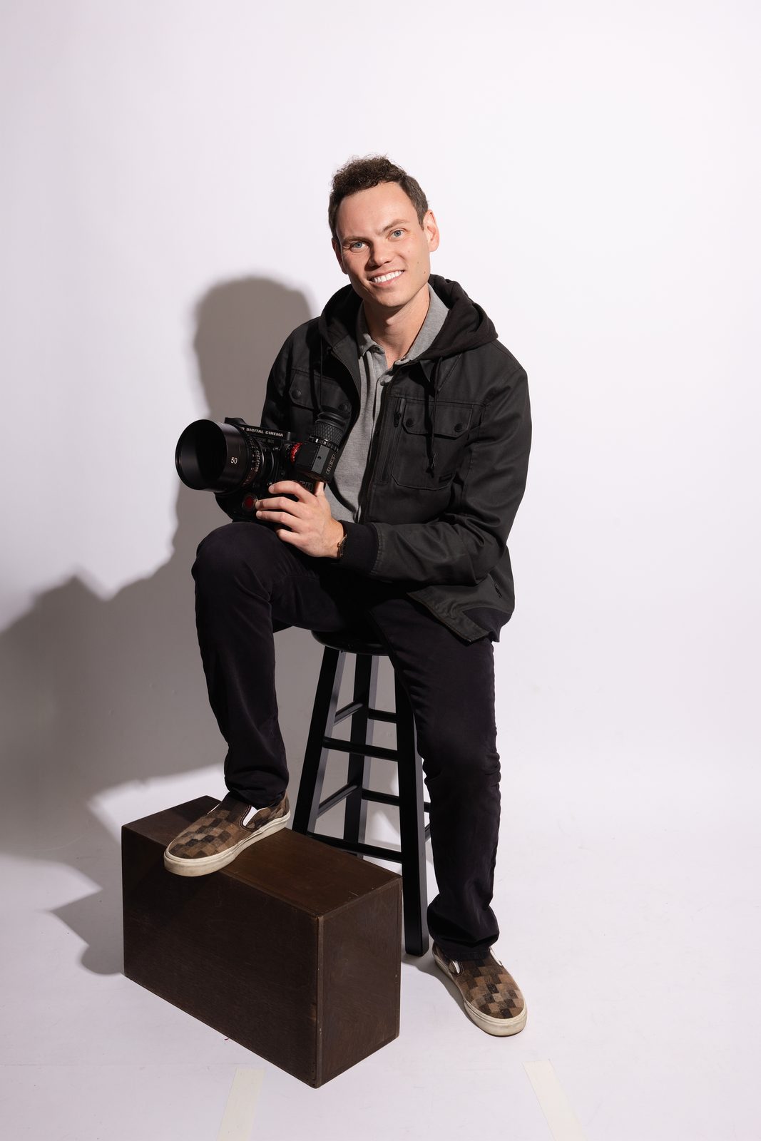 Michael Cecchinelli smiling on a stool with a cinema camera on his knee, in a black hooded jacket against a white studio wall