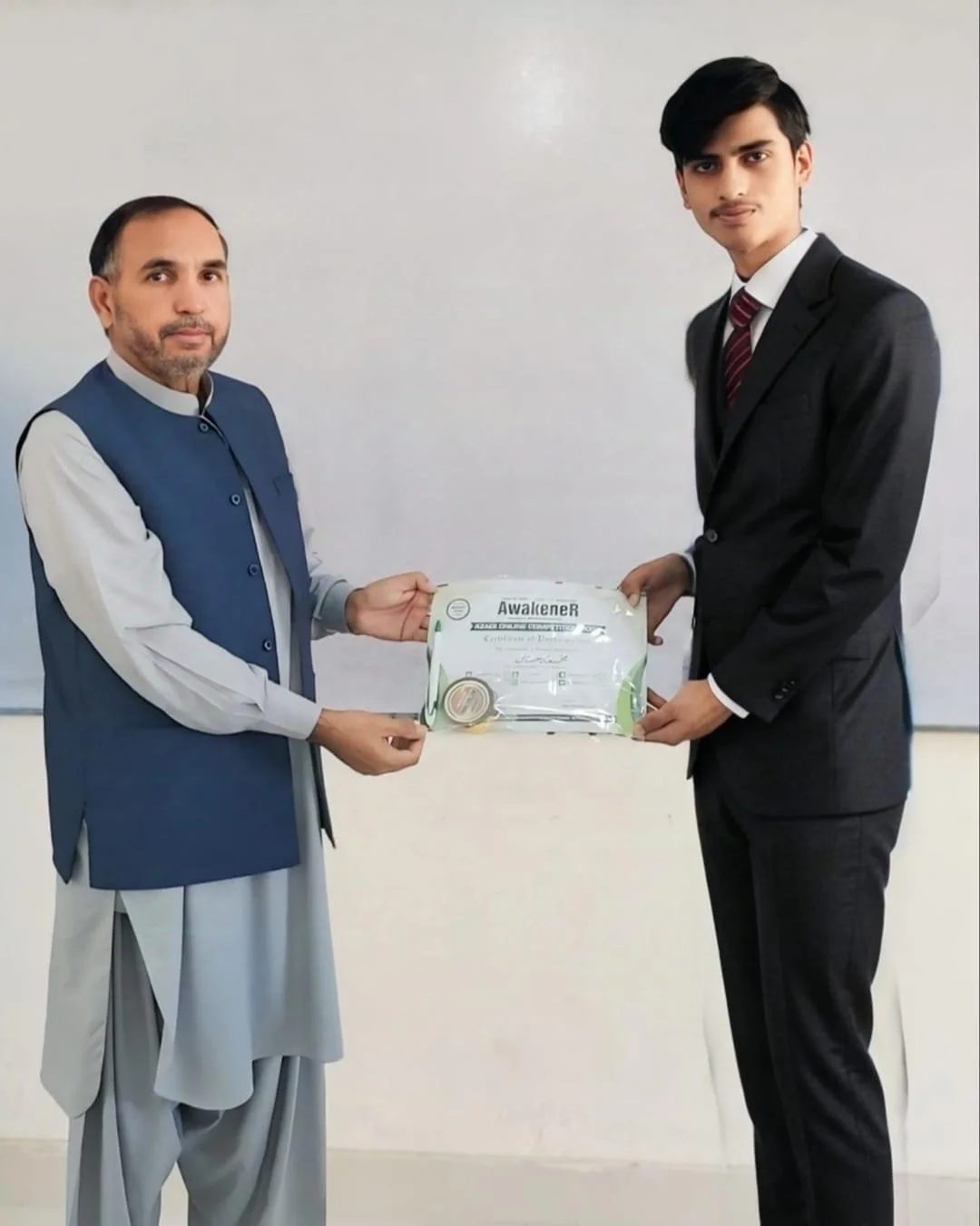 Two men, one in traditional South Asian attire and the other in a suit, exchanging a certificate in a plain room.