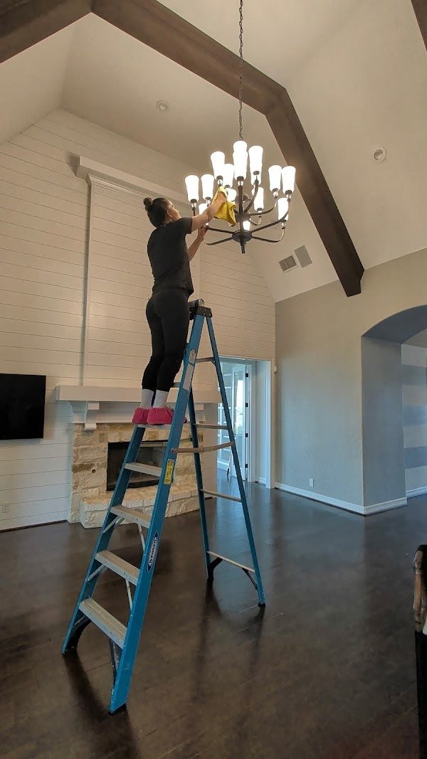 Renee Ambrose cleaning a chandelier on a ladder in a vaulted living room