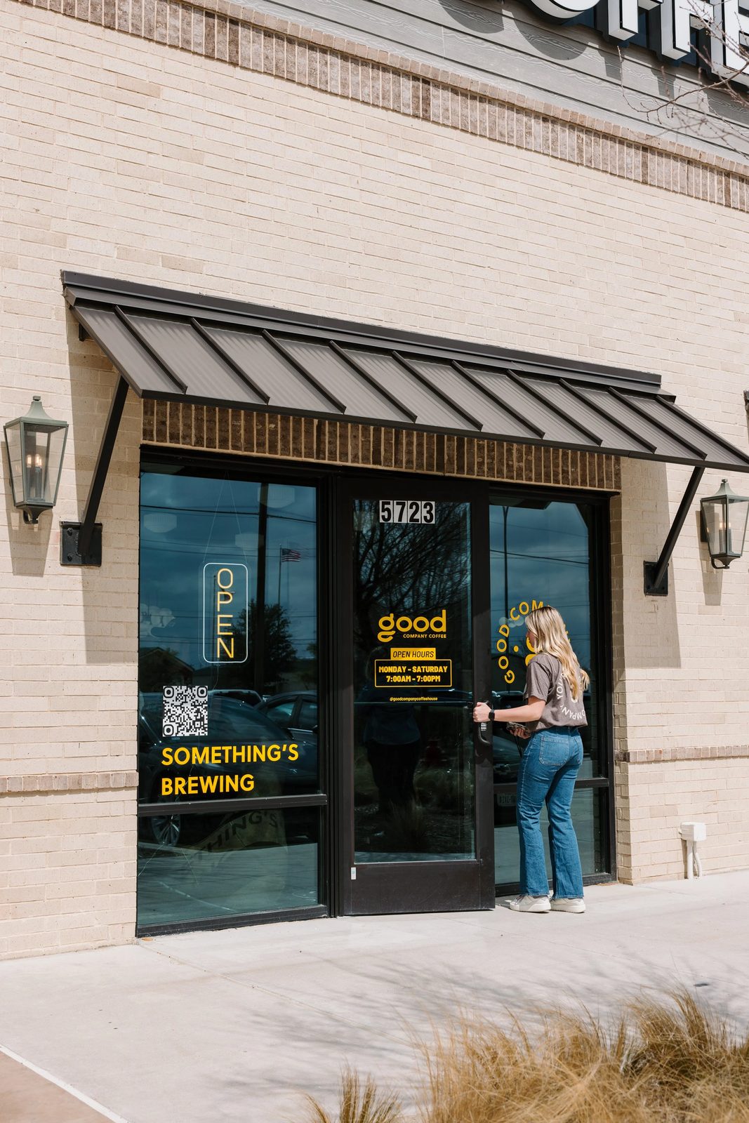 A woman standing at the entrance of a coffee shop with glass doors. The shop has a sign that reads 'good' and offers coffee, with opening hours from 7:00 AM to 7:00 PM Monday through Saturday. The door displays a yellow sign that says 'Something's Brewing' and a QR code. The building has a brick facade with wall-mounted lantern lights.