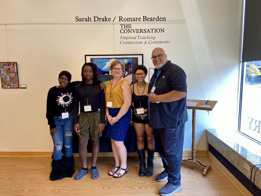 Sarah Drake in a mustard top and blue skirt standing with four guests in front of her collage work under the exhibition title Sarah Drake / Romare Bearden: The Conversation