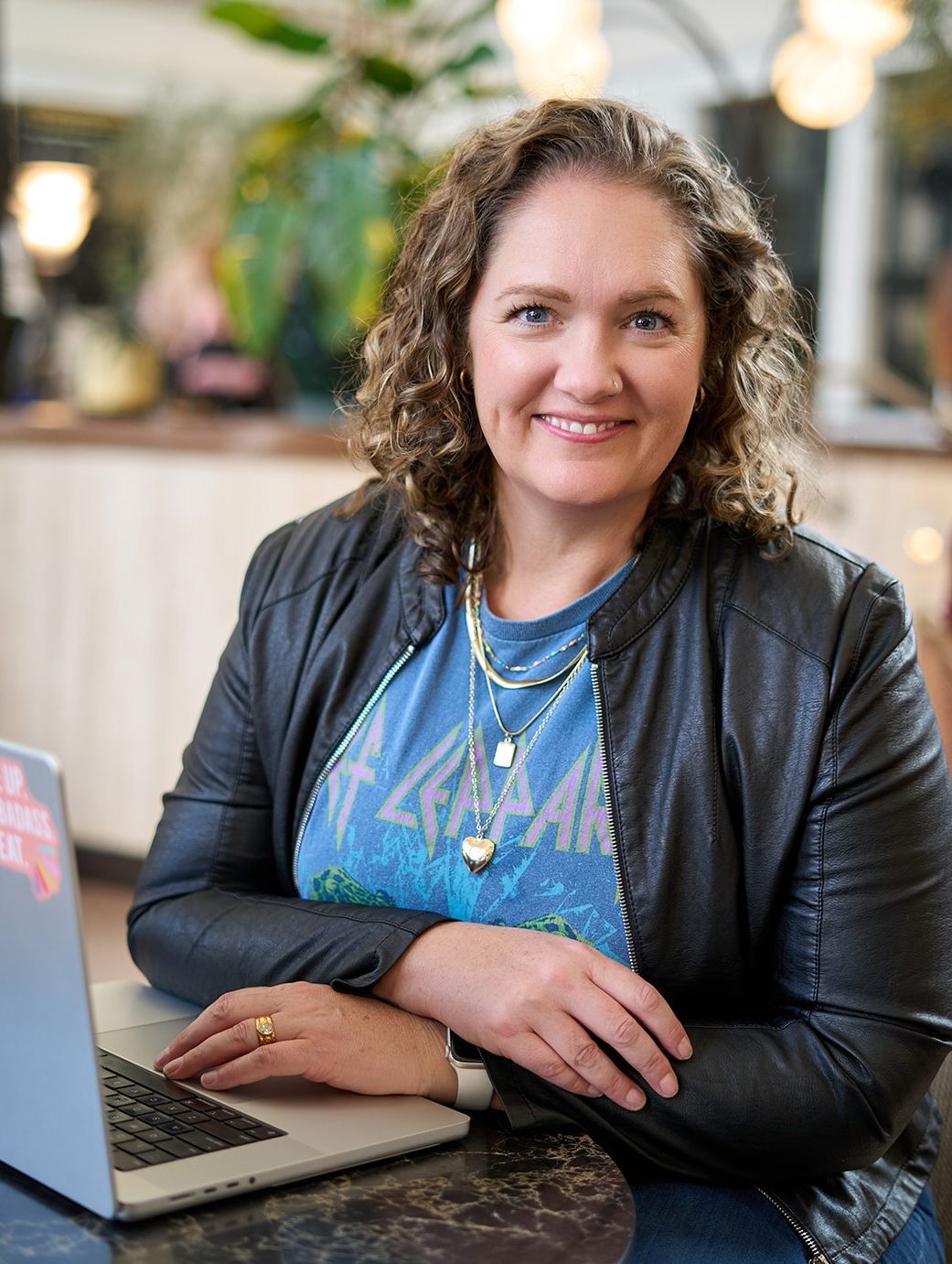 Shannon Gross, founder of Studio One44, smiling at her laptop in a black leather jacket and Def Leppard T-shirt