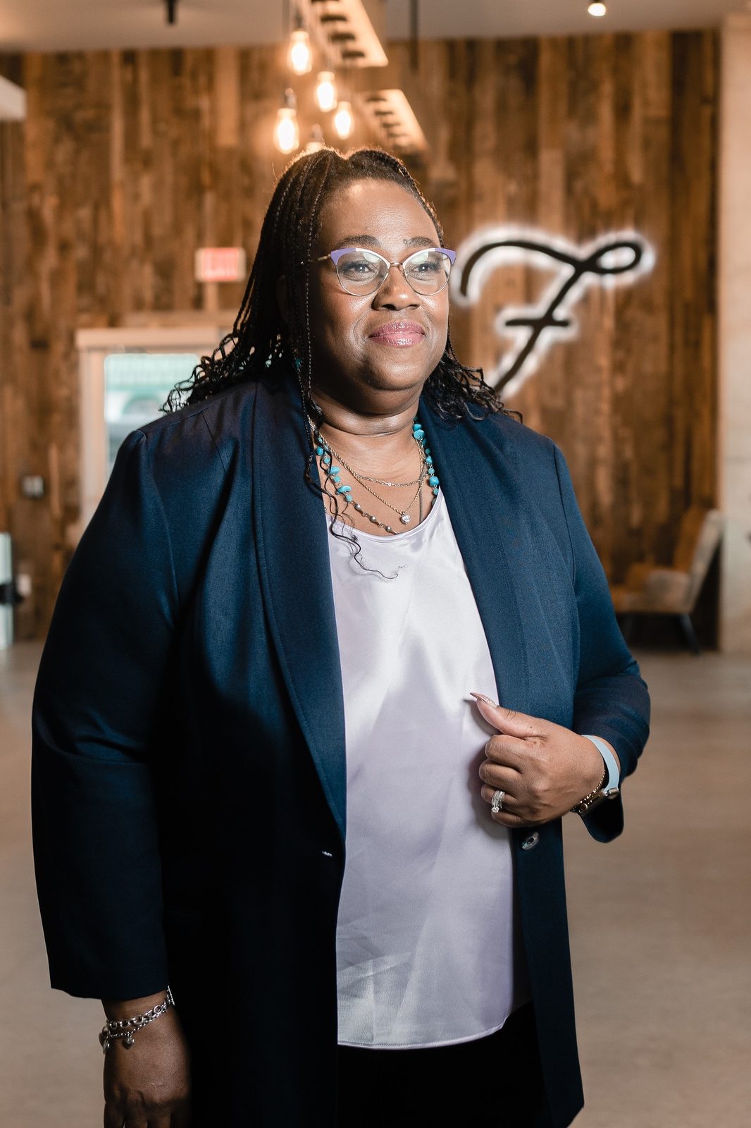 Dr. Sharon M. Jones in a navy blazer and turquoise necklace standing in a lobby with a wood-panelled wall and a glowing script sign
