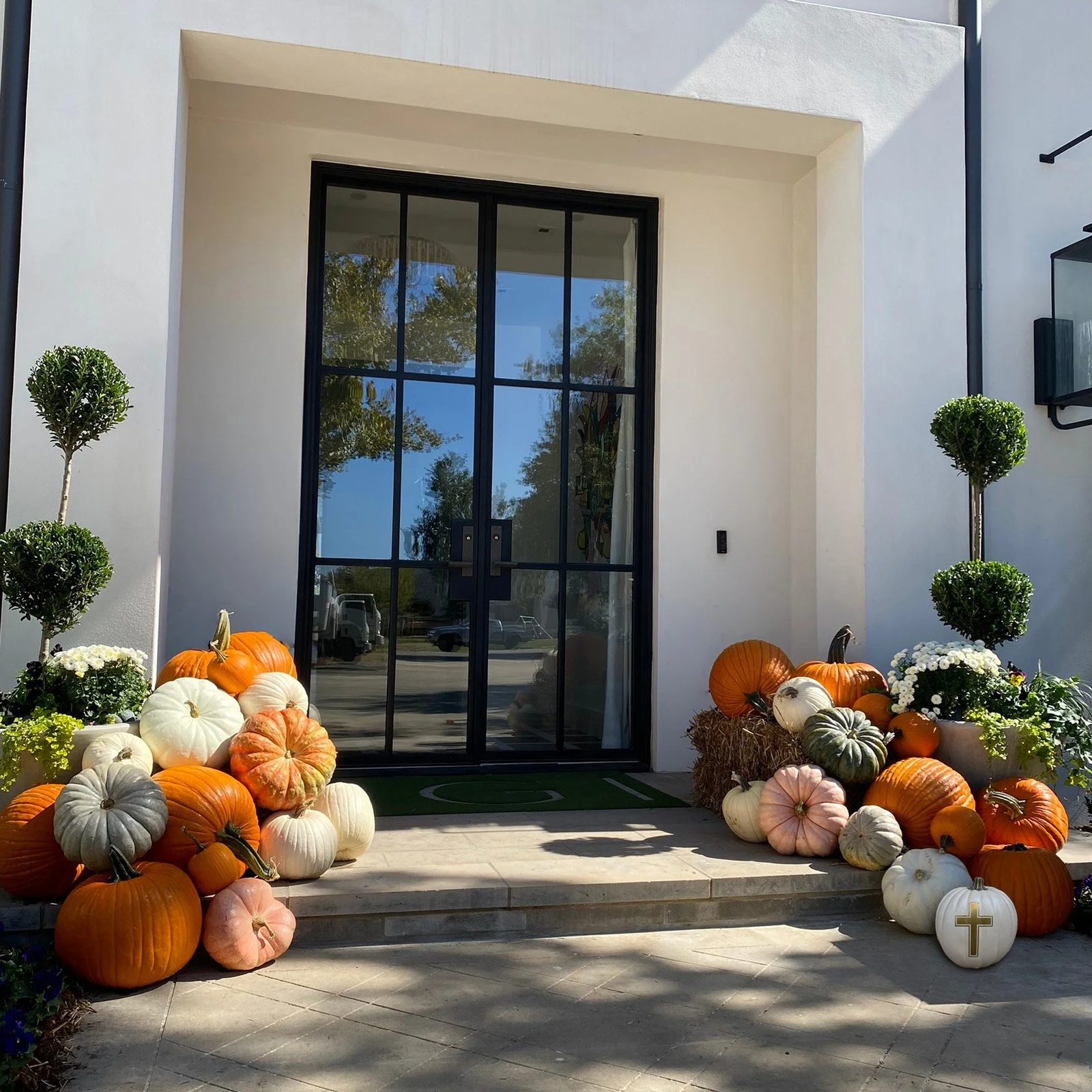 Decorative pumpkins and fall flowers arranged outside a modern white building entrance with a glass door, flanked by small topiary trees.