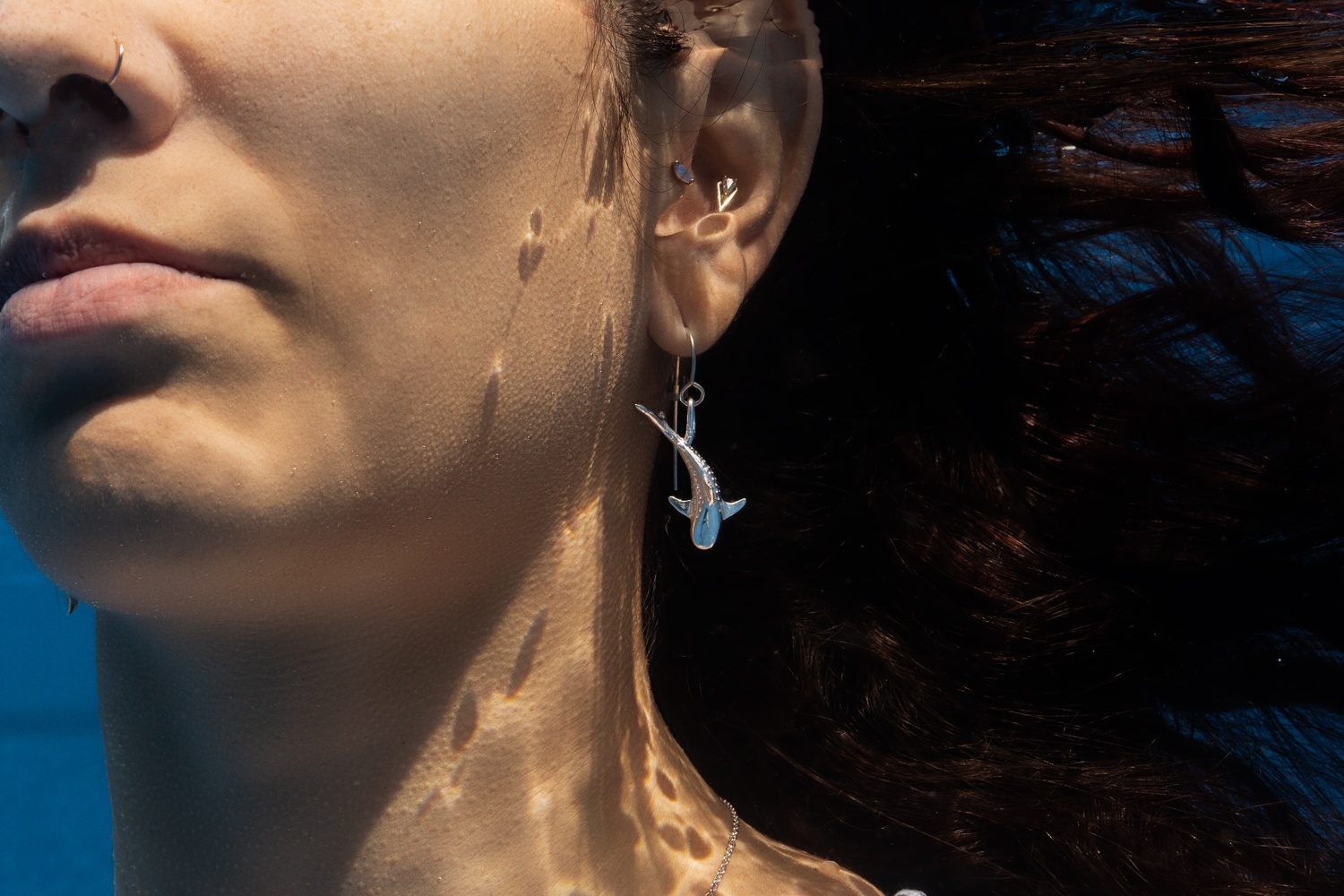 A silver ʻAlohi Kai shark earring worn underwater, hair floating behind