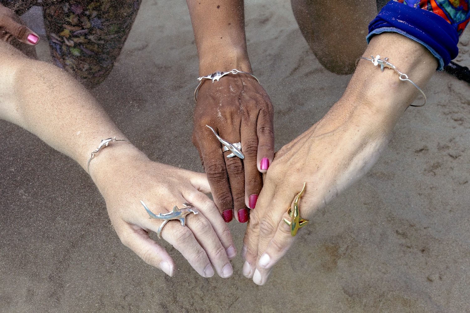 Three pairs of hands on sand wearing ʻAlohi Kai shark and manta rings and bracelets