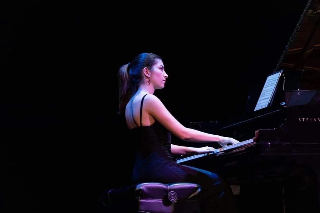 A woman playing a grand piano on a dark stage, wearing a black dress with thin straps.