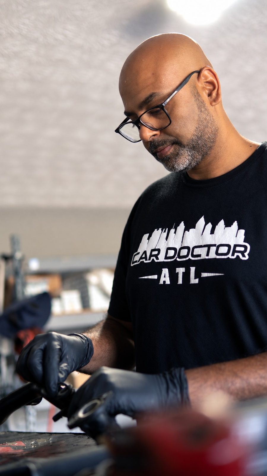 Sri Gadde, founder of CarDoctorATL, in glasses and a Car Doctor ATL t-shirt working on a part in gloves