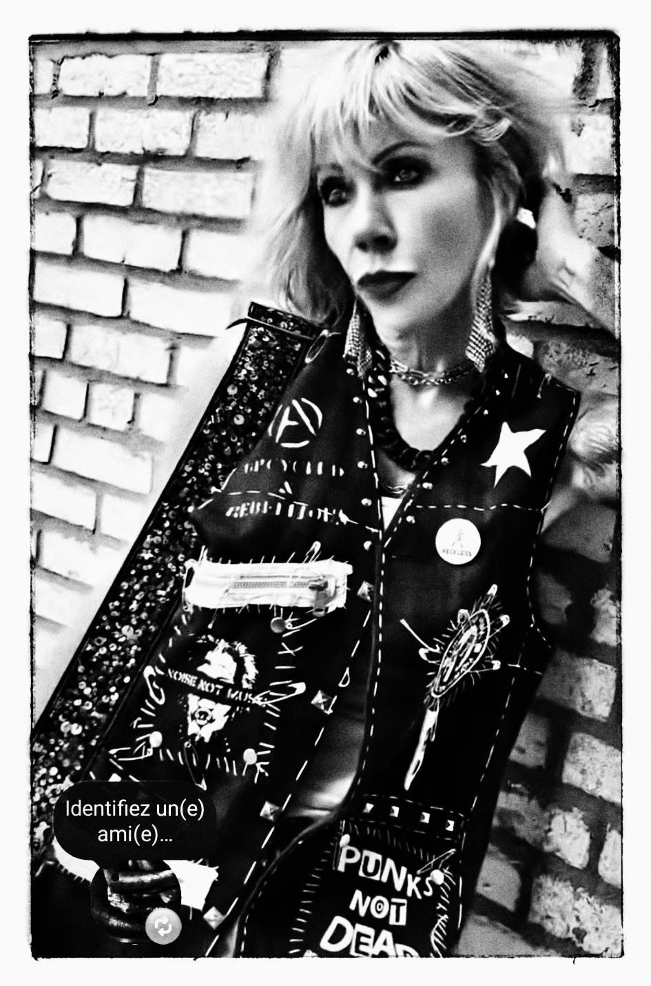 Black and white portrait of Staci Caillé in a studded punk vest against a brick wall