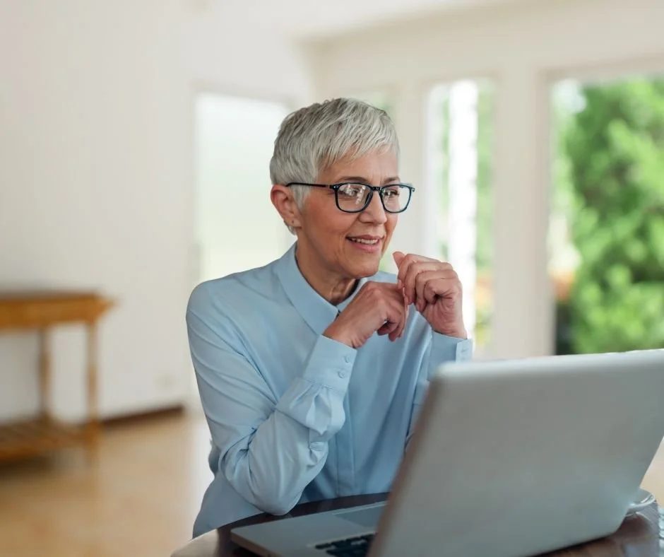 An older woman with short gray hair and glasses smiling while sitting at a table with a laptop in a bright room with large windows and greenery outside.