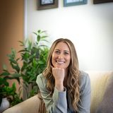 Alexis Flores smiling on a cream sofa in her therapy office, chin resting on her hand, beside a leafy green plant