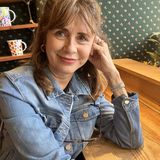 Amy Mink, founder of Amy Mink Design, smiling in a denim jacket at a wooden table in front of green floral wallpaper