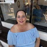 Angie Stewart, owner of Dallas Placenta Encapsulation, smiling in a light blue off-the-shoulder dress outside a storefront in Sanger, Texas