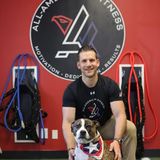 Antonio Somma Jr., founder of All-American Fitness in Morris Plains, New Jersey, kneeling with a dog on the gym's turf