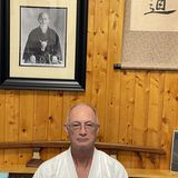 Bill Hague in a white aikido gi, seated in front of a wood-paneled wall beneath a framed portrait of the founder of aikido