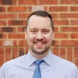 Casey Carmical, founder of 1-FIND SERVICES, smiling in a checked shirt and blue tie in front of a red brick wall