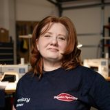Christena Plummer, founder of Plummer Upholstery, in her workshop in Fallston, Pennsylvania, wearing a navy shop shirt with the Plummer Upholstery logo, sewing machines behind her