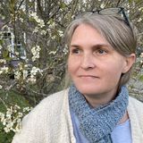 Gabrielle Lazarus, founder of Sacredsong Birth & Wellness, in a cream cardigan and knit scarf beneath a flowering tree