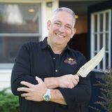 Mike Riddle, founder of Own The Fire, smiling with arms crossed and holding an engraved chef's knife