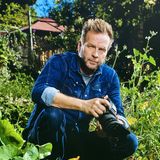 William Sanford, founder of H.I.P Productions, kneeling in a Los Angeles garden in a denim shirt with his camera in hand