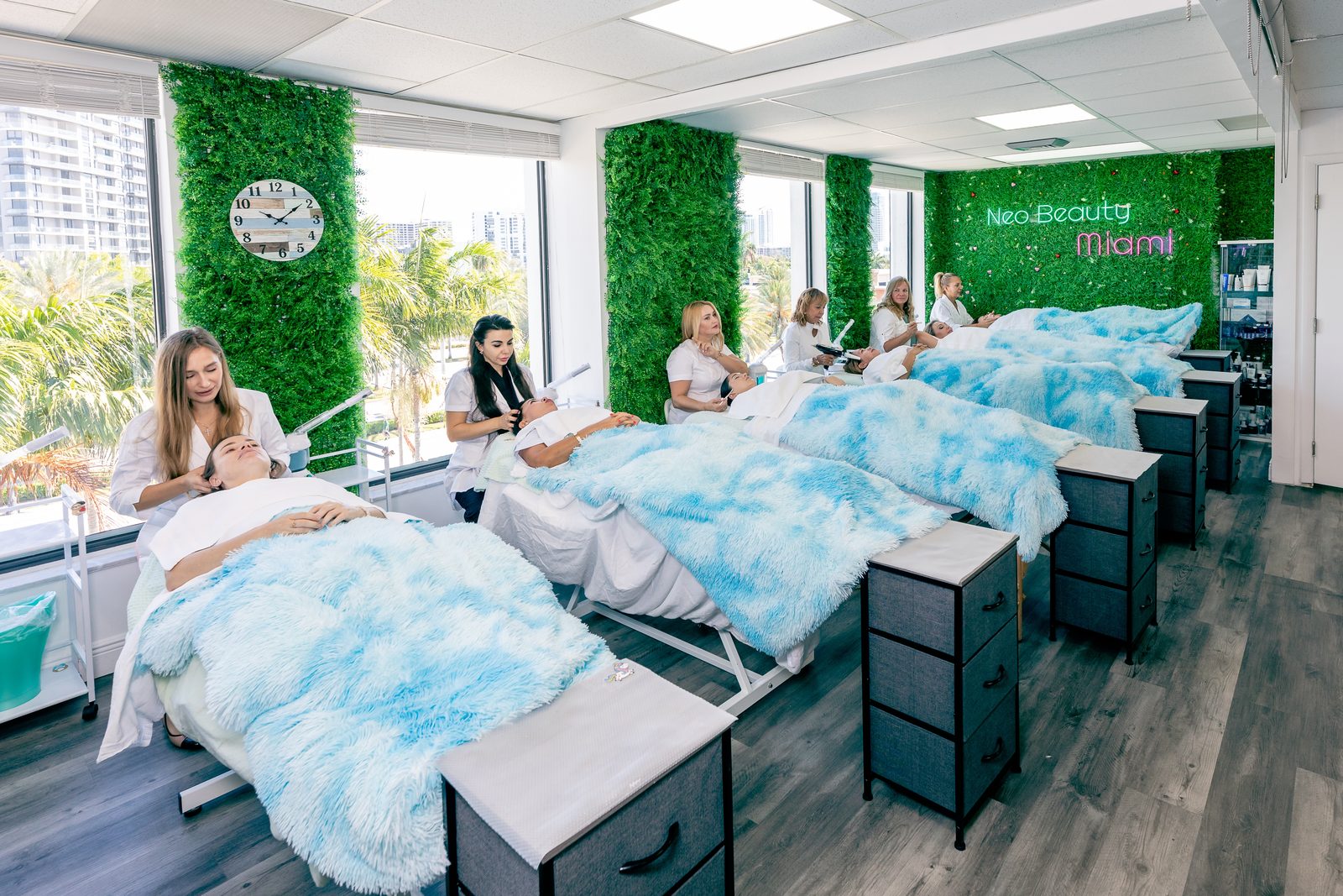 The treatment floor at Neo Beauty Miami, with students giving facials on a row of beds beneath a neon Neo Beauty Miami sign and green walls
