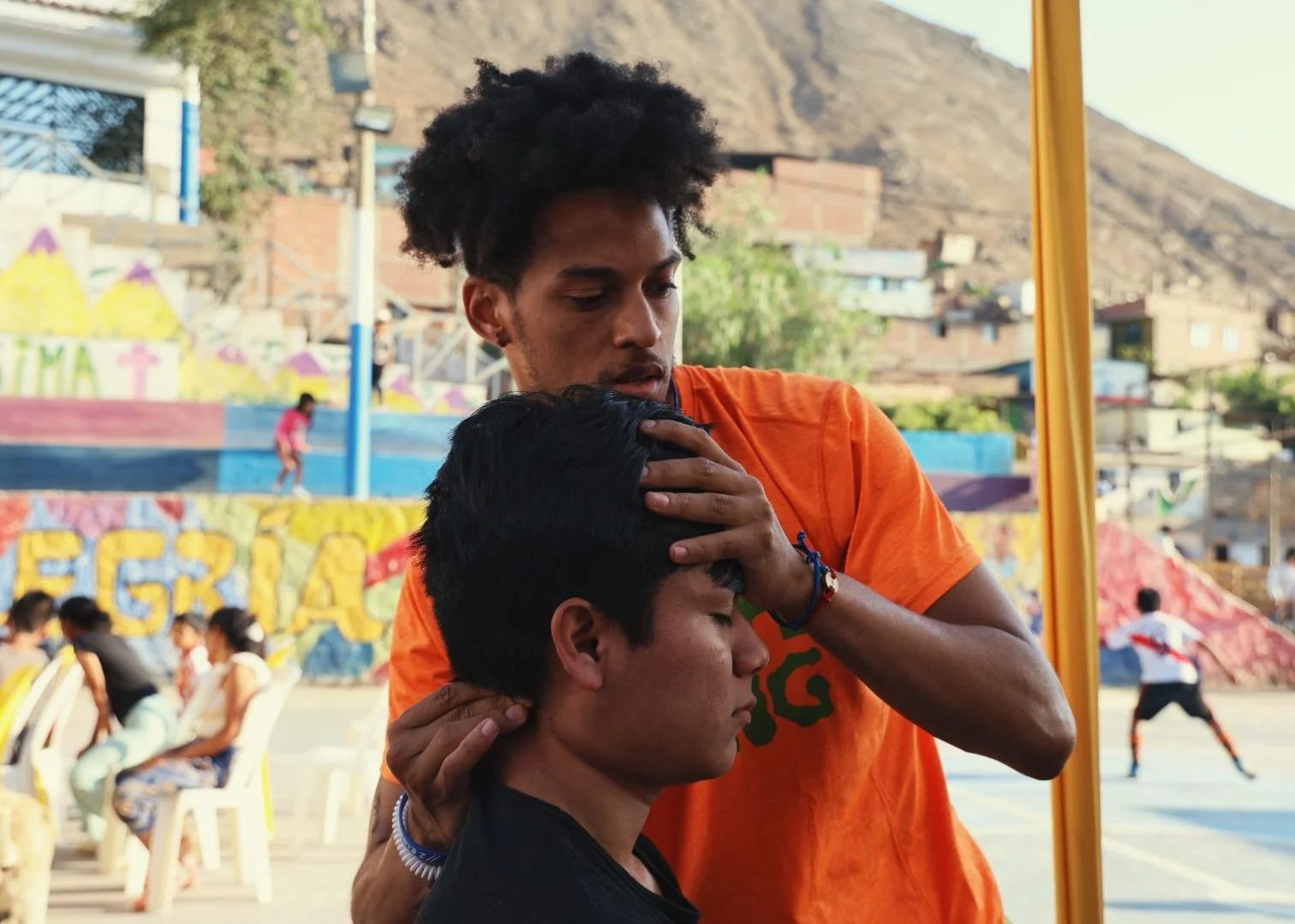 A man in an orange shirt gives a haircut to a young man, outdoors with children playing in the background.