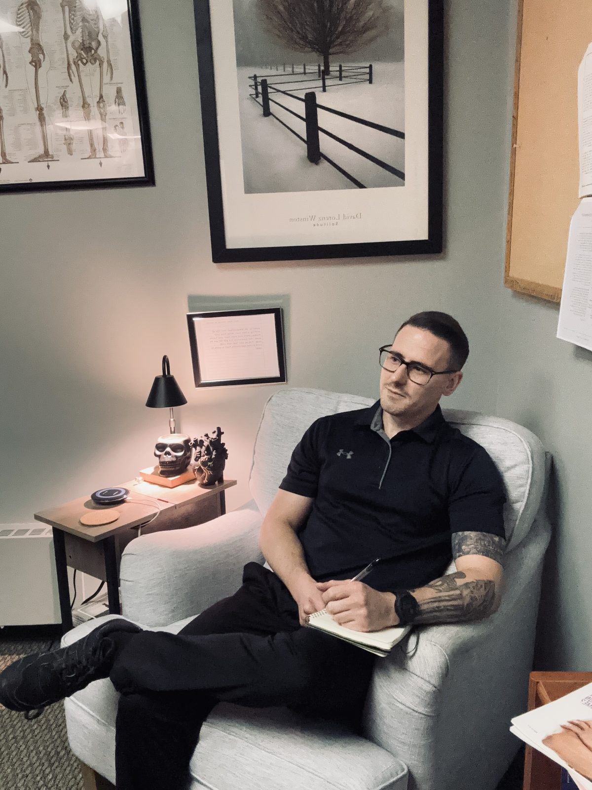 Zach Nuss seated in an armchair in his office, writing in a notebook beneath an anatomy chart